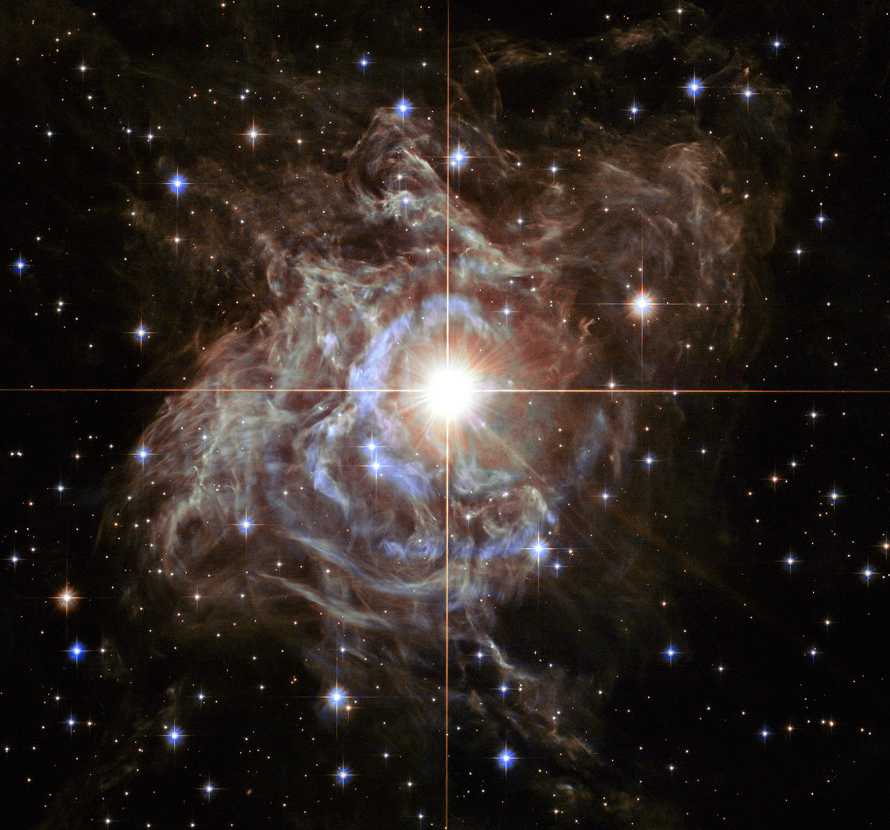 A bright star is at the centre surrounded by blue and white arcs and wisps of dusty circumstellar material. There are several bright white and blue stars across the image which is set against a dark sky. 